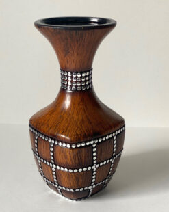 Уникальное сочетание природной эстетики и индустриального стиля. Эта керамическая ваза искусно имитирует фактуру натурального дерева с глубоким теплым оттенком и выразительным рисунком волокон. Особый характер изделию придаёт декор в виде металлических заклепок, которые образуют строгий геометрический узор и эффектно подчеркивают изгибы формы. Контраст «деревянной» матовой поверхности и холодного блеска металла делает вазу самодостаточным арт-объектом. Она идеально дополнит интерьеры в стиле лофт, эко-минимализм или современное кантри. Благодаря устойчивому основанию и оригинальному расширяющемуся горлышку, ваза прекрасно подходит для флористических композиций из живых цветов или сухоцветов. Характеристики: Материал: керамика с декоративным покрытием. Текстура: имитация натурального дерева, металлические элементы. Цвет: орех / хром. Стиль: лофт, эко, современный.